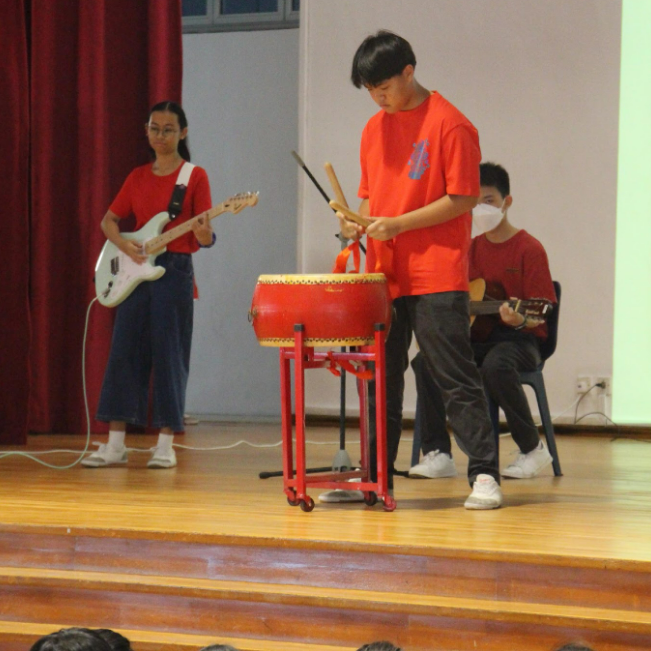 A student in orange strikes a red Chinese drum on stage while another holds a white electric guitar before a seated audience.